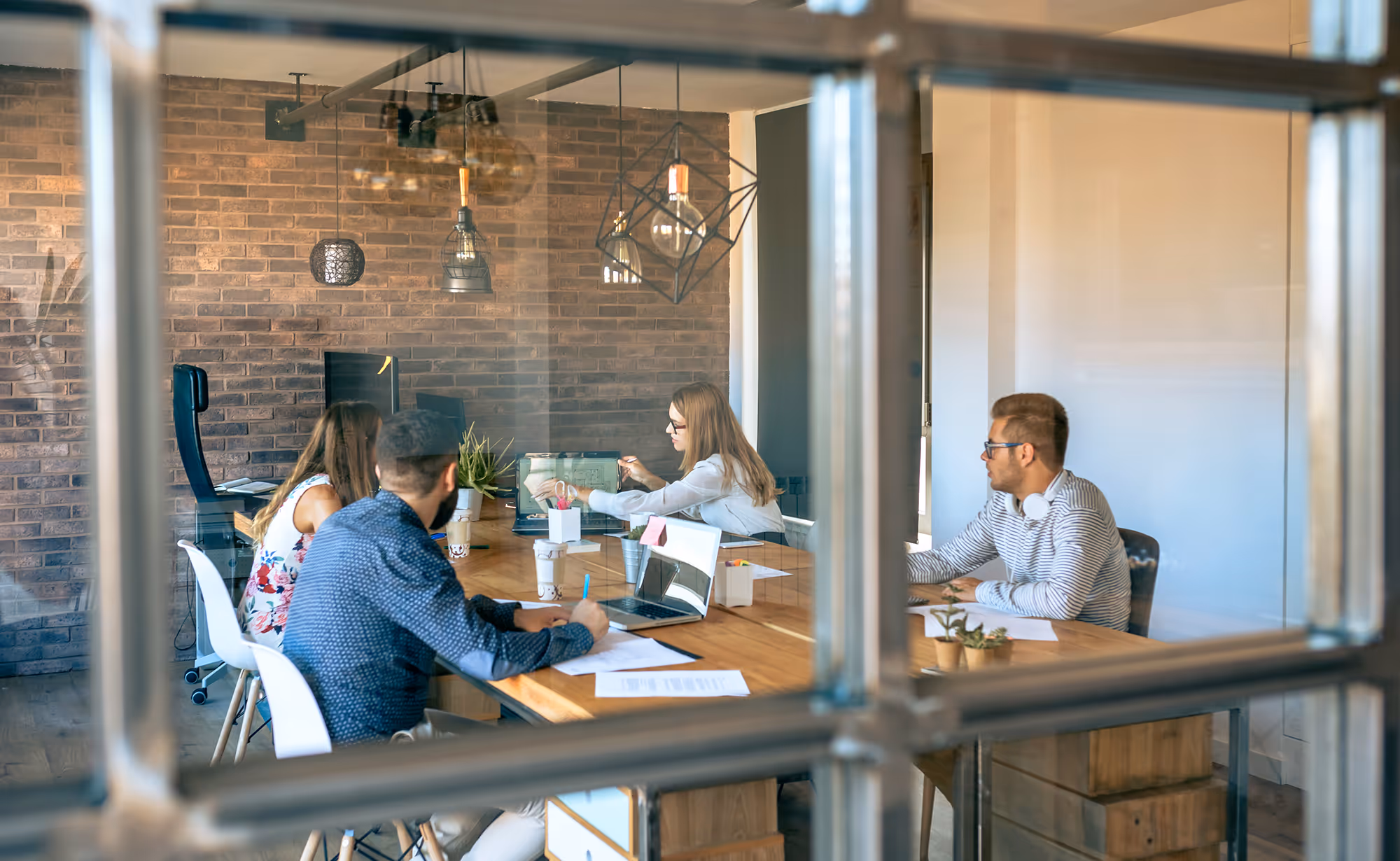 Vier jonge volwassenen werken samen aan een houten tafel in een moderne kantoorruimte met bakstenen muren en hanglampen.